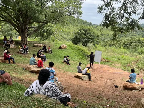 Schoolchildren sitting on rocks under the trees for an outdoor class